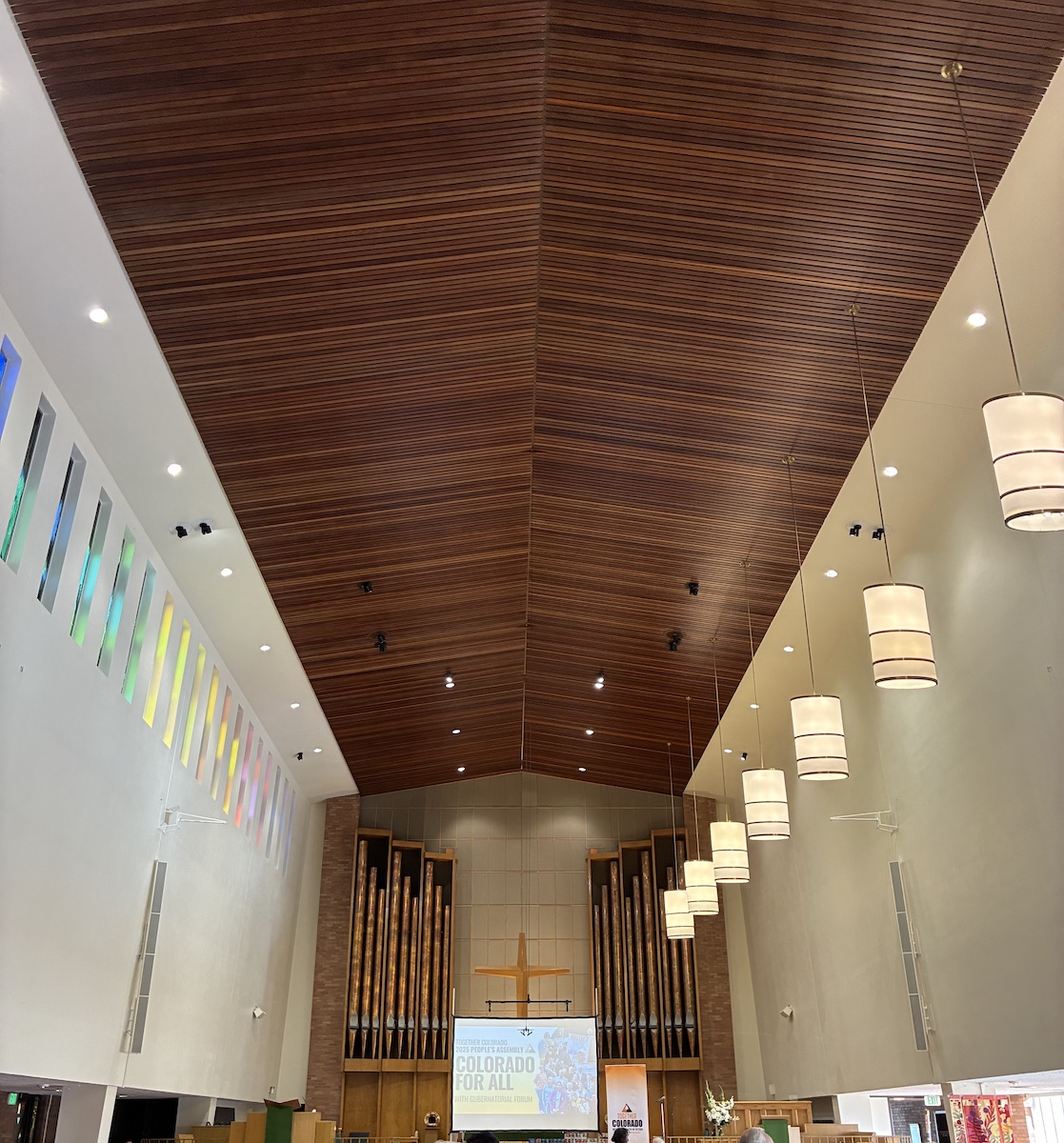 View of the long, tall church interior. A projector screen is set up at the center of the raised platform. Lecterns exist on either side. Pipes at the back. A star-like Christian cross is peaking up behind the projector screen.