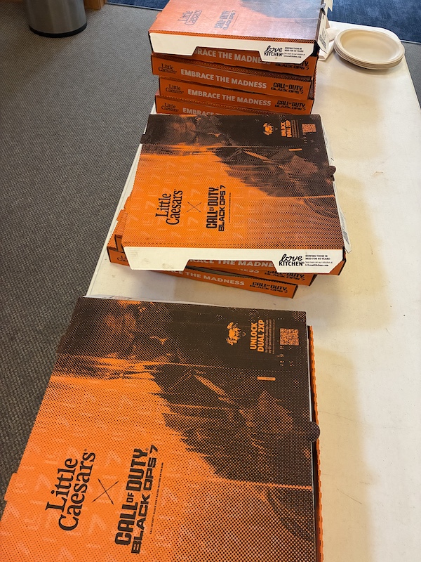 Several identical pizza boxes on a long table sporting Call of Duty Black Ops 7 branding, and the slogan 'Embrace the Madness'.
