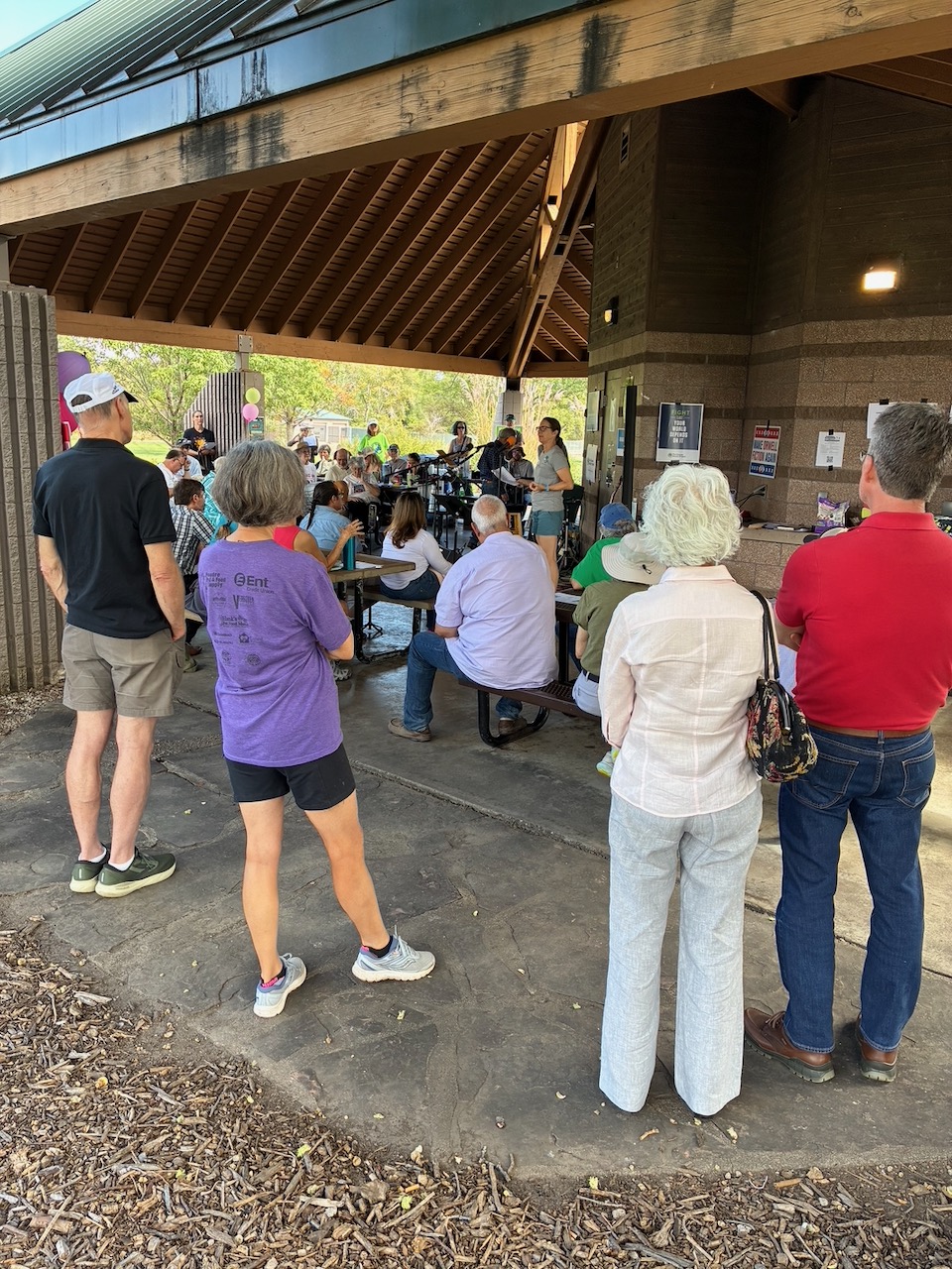 Several locals stand under the covered meeting space at the park, listening to someone speaking. Most are seated.