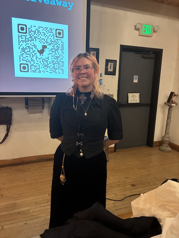 Chris Pruneski stands in her hand-made Victorian-inspired costuming, with hands behind her back. Her glasses have a long chain descending by her face. There is a large broach at her high-necked collar, a small gold amulet, and a pocket watch hangs over her long dark skirt. Her outer layer is fastened by two dark buckles at her middle.