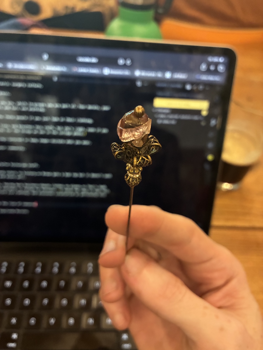 A vintage coppery hairpin with a slightly rosy stone pierced at the end. Small metalwork folds in on itself, the full shape perhaps conjuring an image of a flower, atop the long thin stake. This photo has my hand in it, and behind it is a blurry view of the iPad used to take these notes.