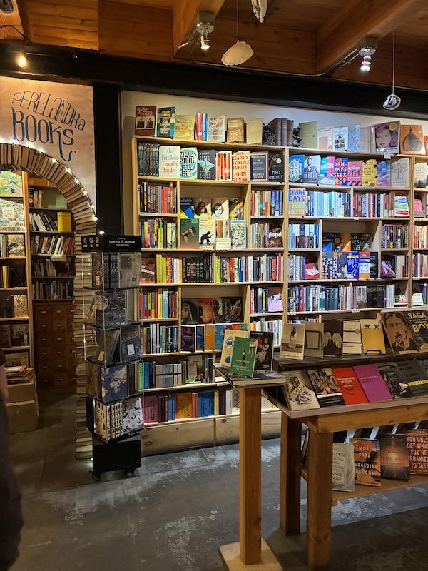 A warm interior of a bookshelf on the wall, an arched path behind to more, and an isle stand with yet more books.